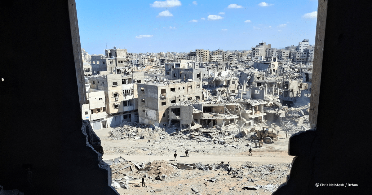 Wide view through a ruined building shows a bombed urban landscape with collapsed apartment blocks, piles of rubble, and a few people near a heavy vehicle in the foreground.