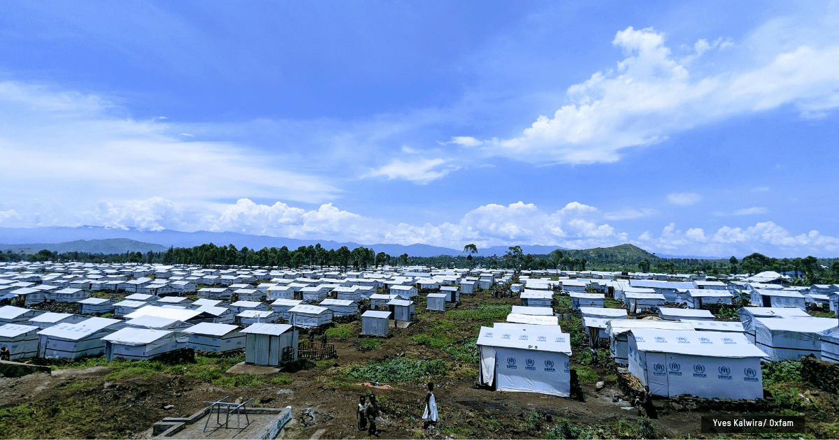 A vast refugee camp with hundreds of white tents spread across a dry field under a clear blue sky; a few people walk in the foreground.