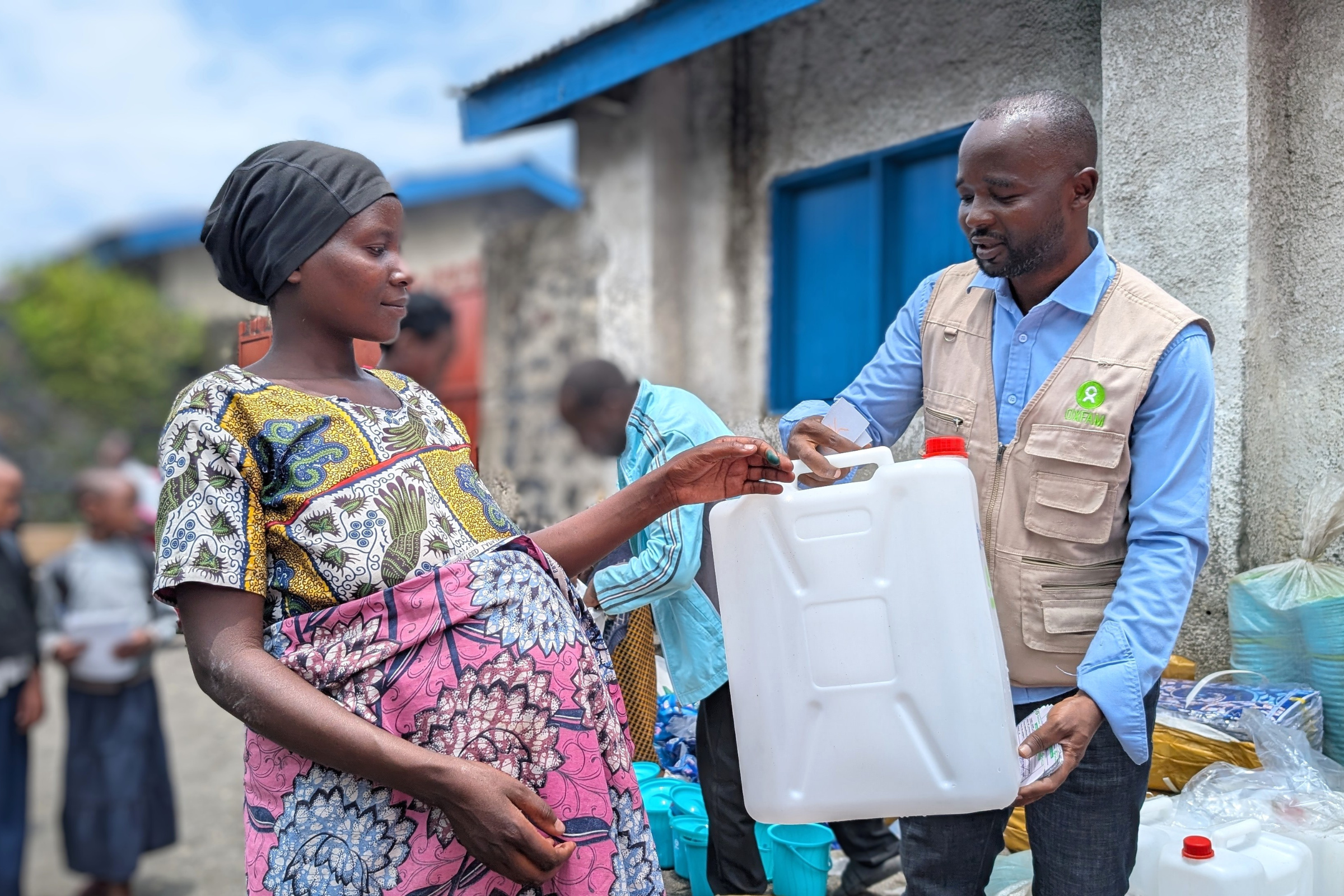 Woman in a colorful dress hands a large white plastic container to a man in a beige vest at an aid distribution site.